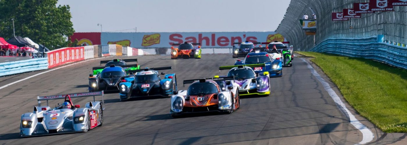 Historic race cars tightly packed on track at Watkins Glen International, speeding side by side under a Sahlen’s billboard during a sports car race.