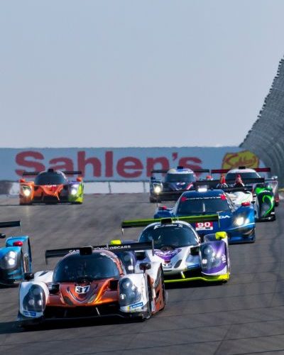 Historic race cars tightly packed on track at Watkins Glen International, speeding side by side under a Sahlen’s billboard during a sports car race.
