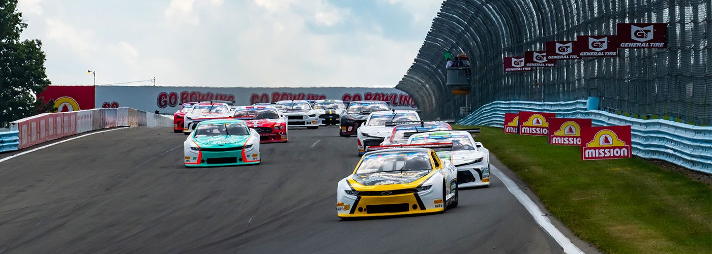 A pack of stock cars races side by side on a paved track at Watkins Glen International. The lead car is yellow and white, followed closely by cars in teal, red, black, and white. The cars are heading uphill toward a curve, with safety fencing and sponsor signs along the right side of the track. The sky is partly cloudy overhead.