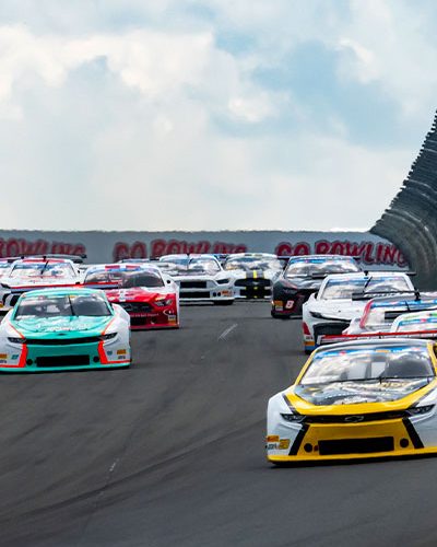A pack of stock cars races side by side on a paved track at Watkins Glen International. The lead car is yellow and white, followed closely by cars in teal, red, black, and white. The cars are heading uphill toward a curve, with safety fencing and sponsor signs along the right side of the track. The sky is partly cloudy overhead.