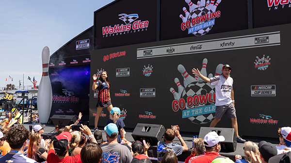 Hosts on a stage at Watkins Glen International lead a fan interaction during a “Go Bowling at The Glen” event, with a cheering crowd gathered in front of the stage.