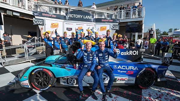 Acura Motorsports team celebrates in Victory Lane at Watkins Glen International, posing with their race car and trophies as confetti falls and crew members cheer behind them.