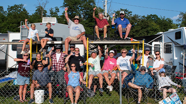 A large group of race fans gathered at a campground, posing together on and around a raised platform behind a chain-link fence. Many are smiling, waving, and holding drinks, with RVs and trees visible in the background under a clear blue sky.