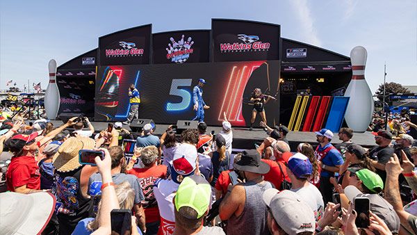 A crowd of fans stands in front of a large Watkins Glen-branded stage featuring a digital screen with a driver and the number 5 displayed. People in the audience are holding up phones to take photos, while presenters or performers stand on stage during a daytime event.