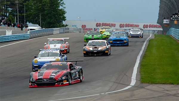 Multiple race cars competing on a road course at Watkins Glen International, led by a red, black, and silver Ford Mustang race car in the foreground. Several brightly colored sports cars follow closely behind on the paved track, with grassy infield areas and safety barriers lining the course. “Go Bowling” signage is visible above the track in the distance under an overcast sky.