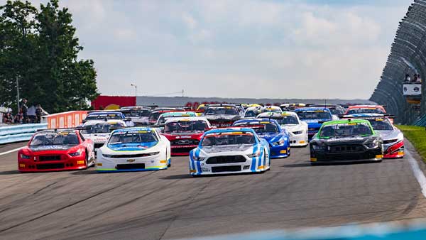 A pack of stock cars races side by side on the frontstretch at Watkins Glen International during a vintage racing event, with multiple brightly colored cars charging toward the camera under partly cloudy skies.