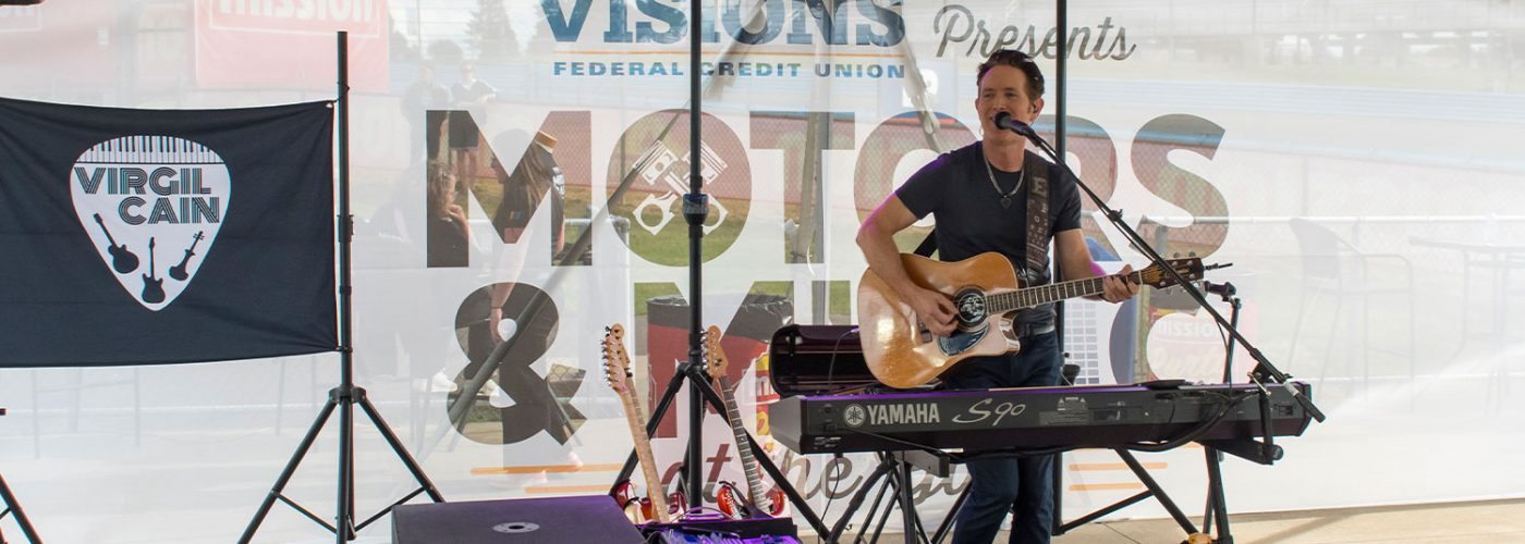 Musician performs with an acoustic guitar behind a keyboard onstage at the Motors & Music event.