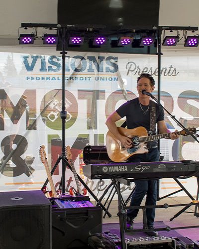 Musician performs with an acoustic guitar behind a keyboard onstage at the Motors & Music event.